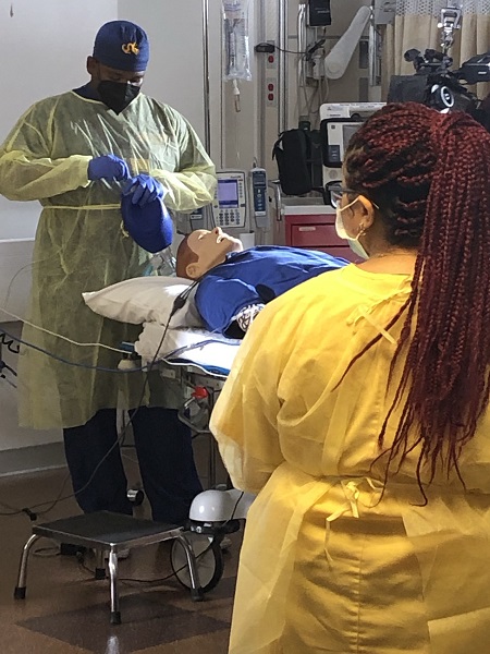 Black male in scrubs and a gown preparing to use ventilation bag on a manikin while another nurse looks on.